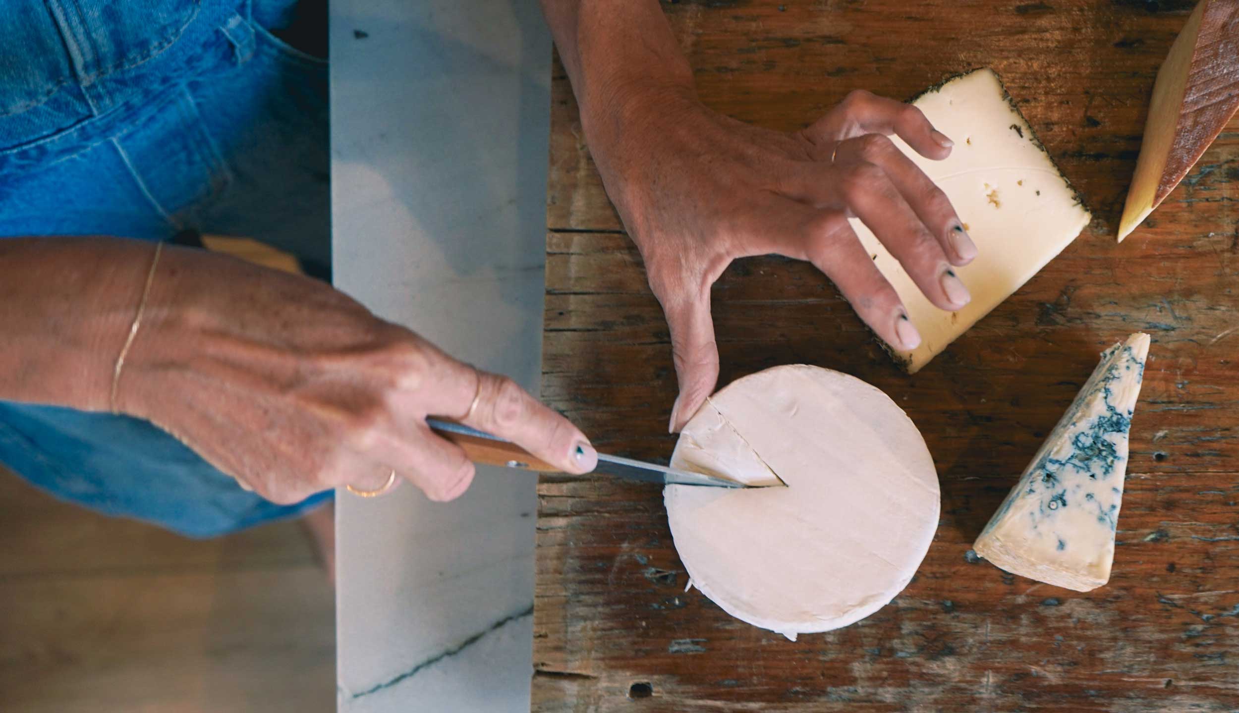 Person cutting a wheel of cheese with a knife on a wooden table.