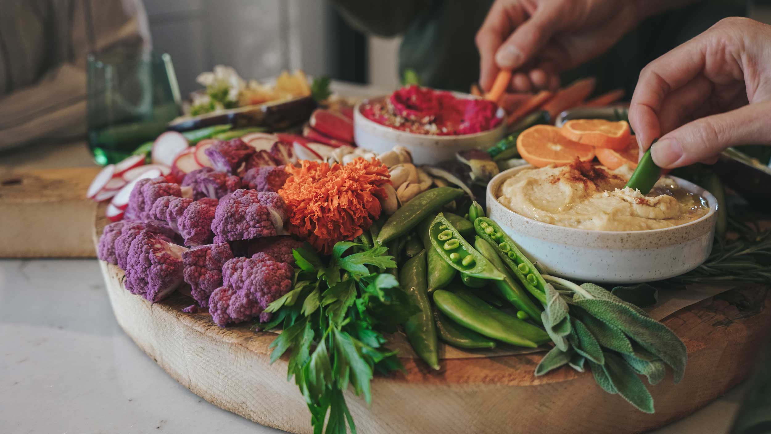 Platter of colorful vegetables and dips with a person dipping a vegetable into a bowl of hummus.