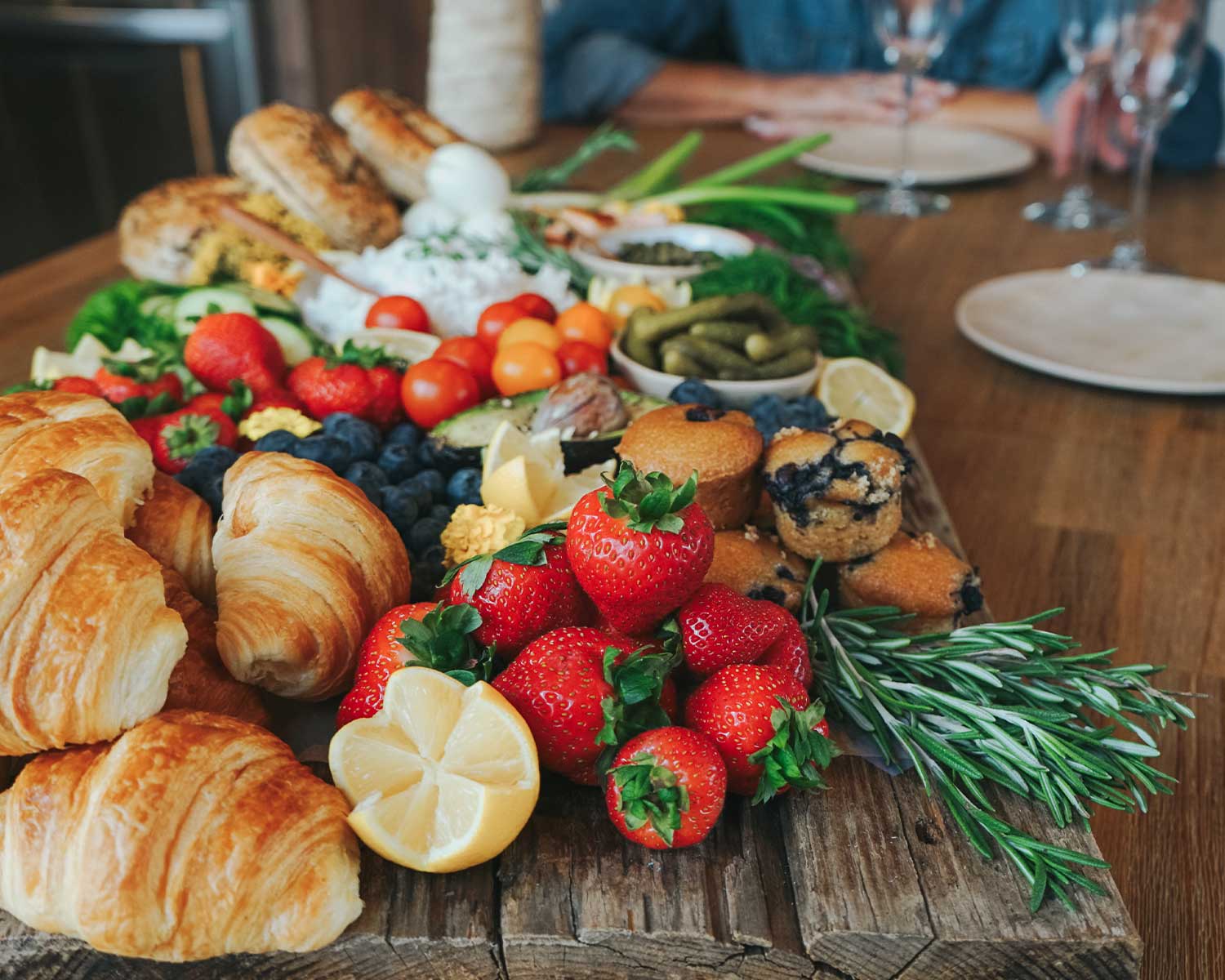 Assorted pastries and fruits on a wooden board with a blurred background of people sitting at a table.