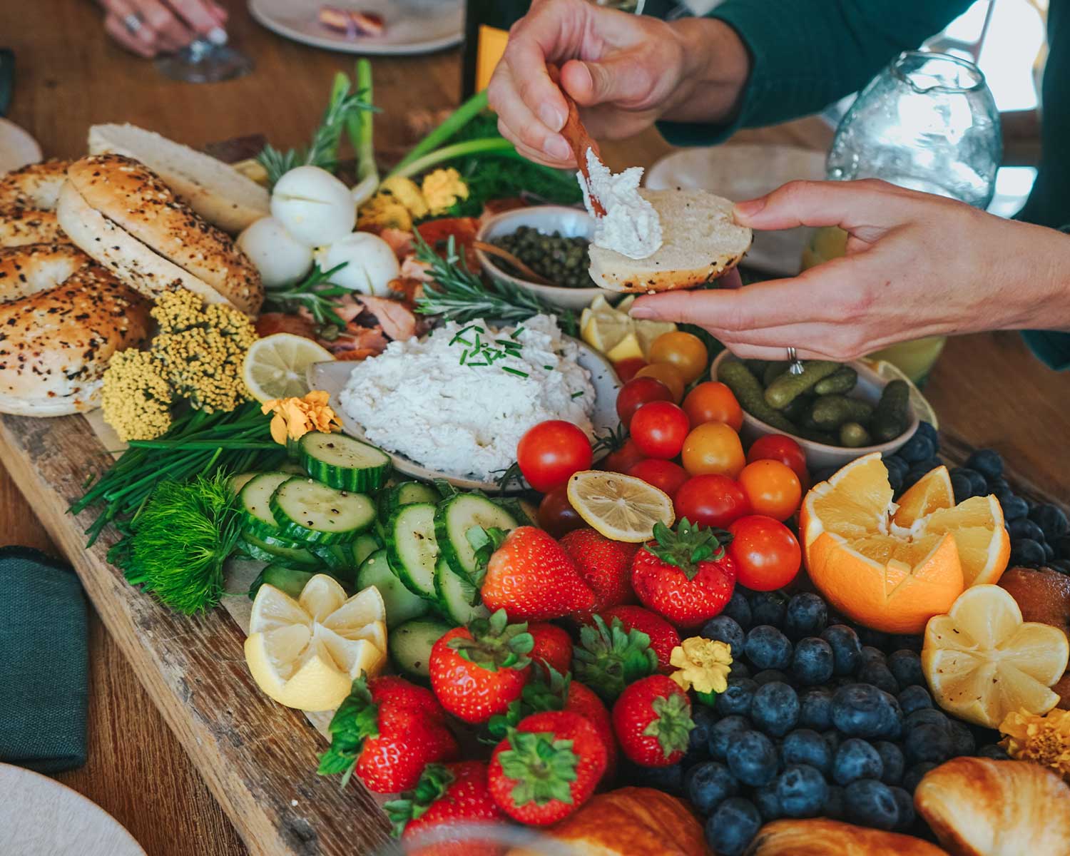 Person preparing a sandwich with a platter of fresh fruits, vegetables, and bread on a wooden table.