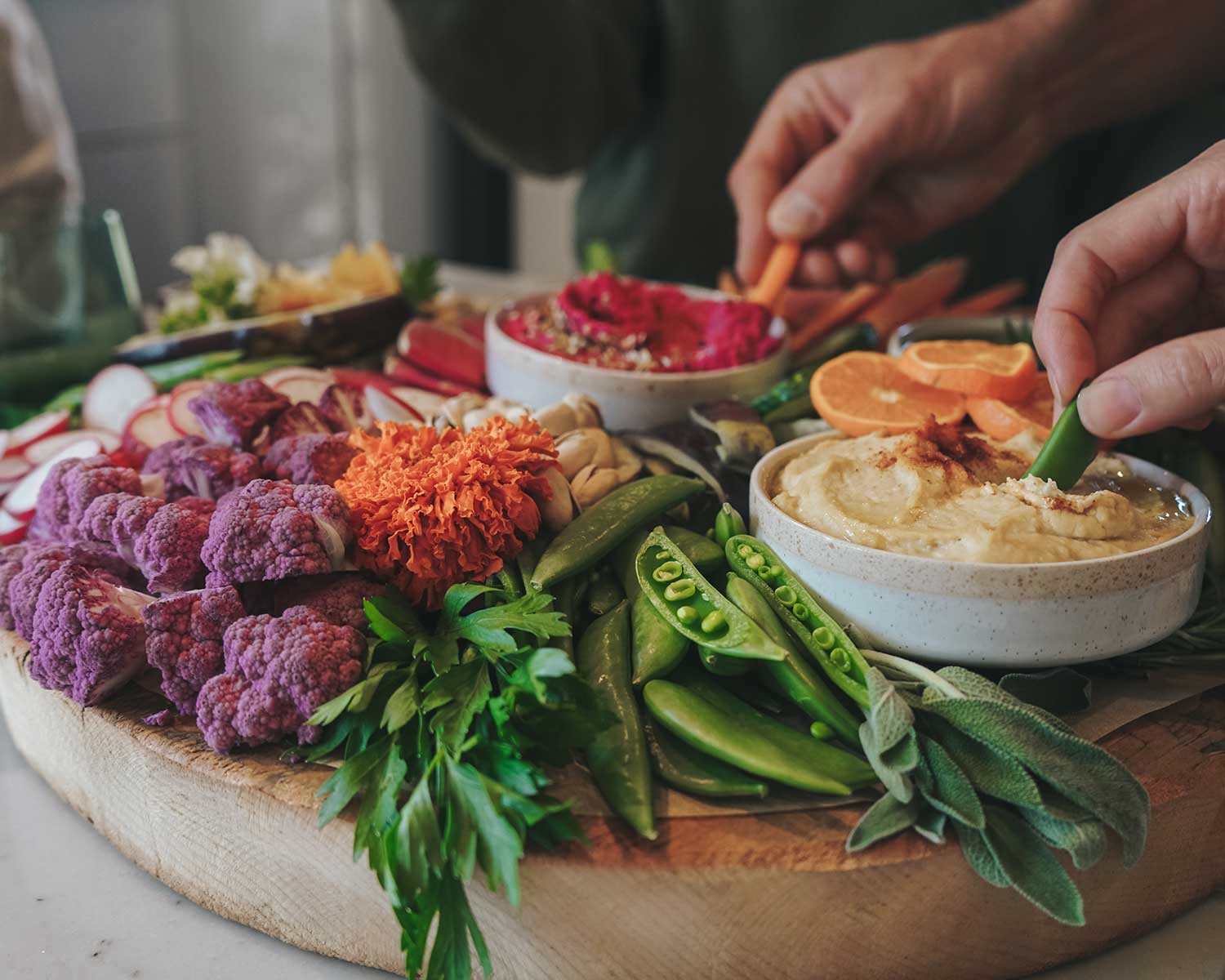 Assorted colorful vegetables and dips on a wooden board with a person dipping a vegetable into a dip.