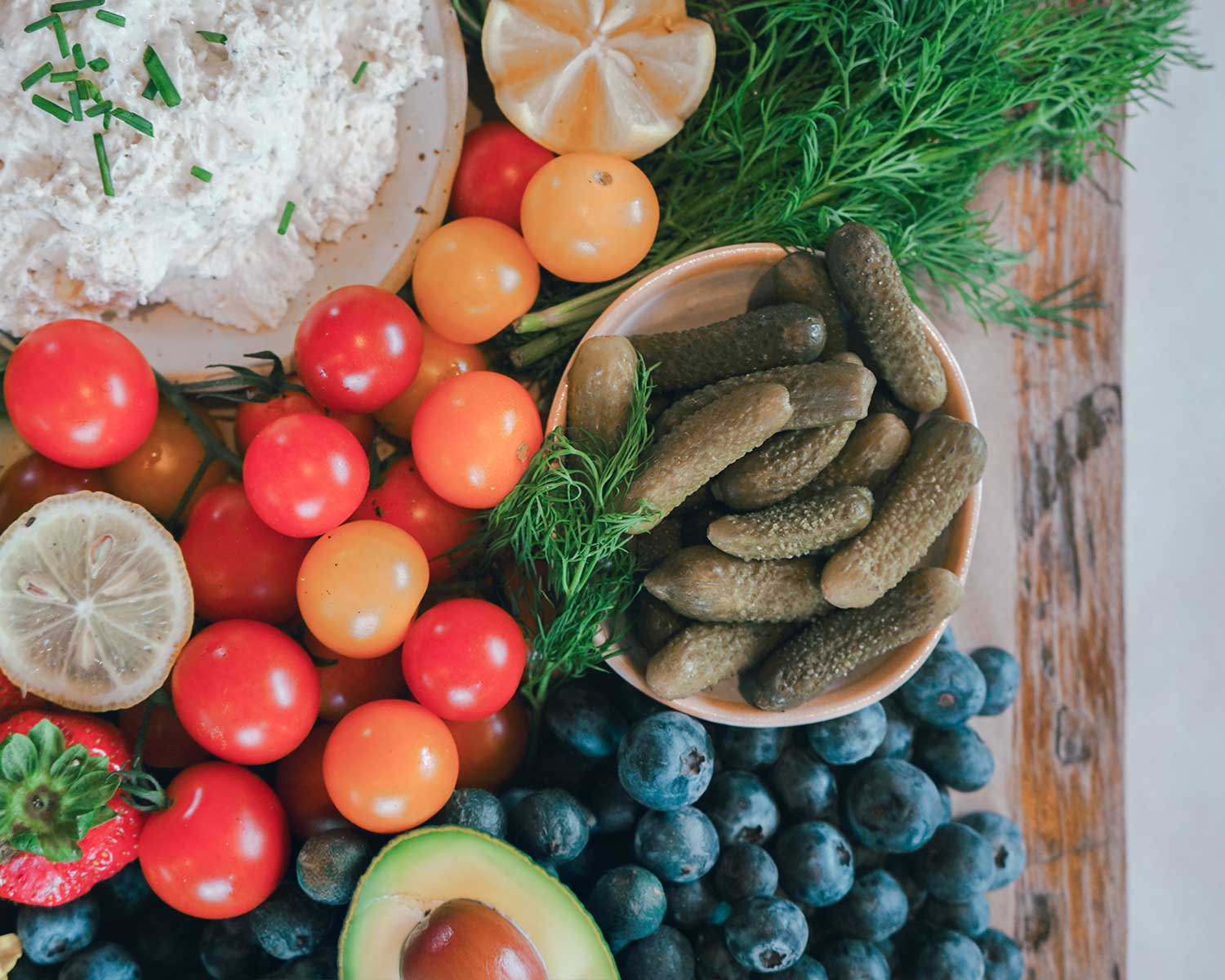Assorted fresh vegetables including tomatoes, blueberries, and pickles on a wooden surface.