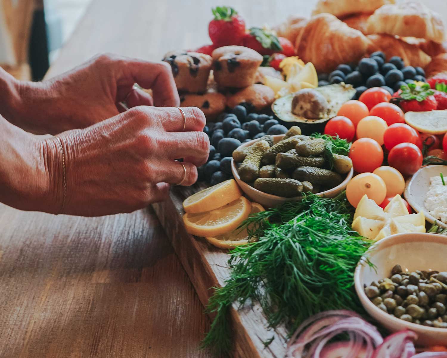 Person preparing a charcuterie board with various fruits, vegetables, and meats on a wooden surface.