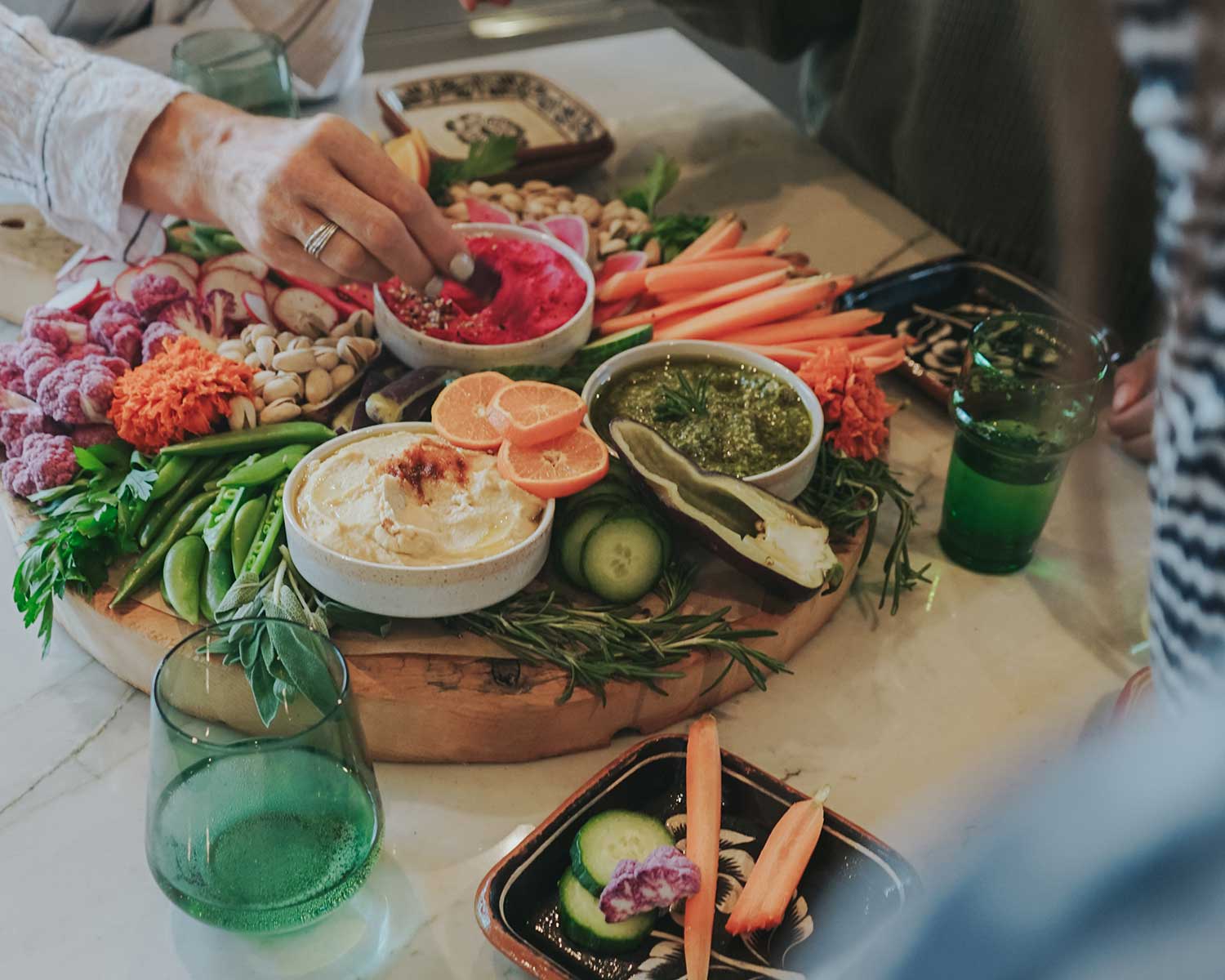 Platter of hummus, vegetables, and dips on a table with people around.