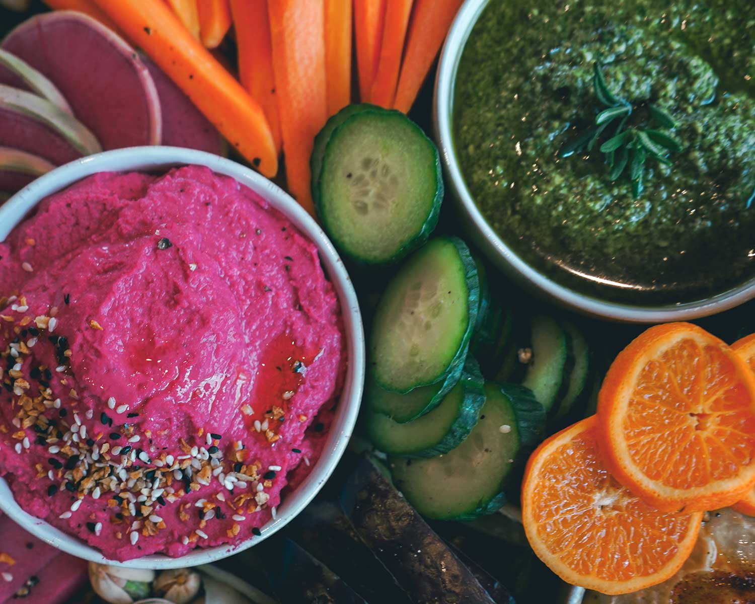 Assorted vegetable platter with pink hummus, green pesto, and sliced oranges on a wooden board.